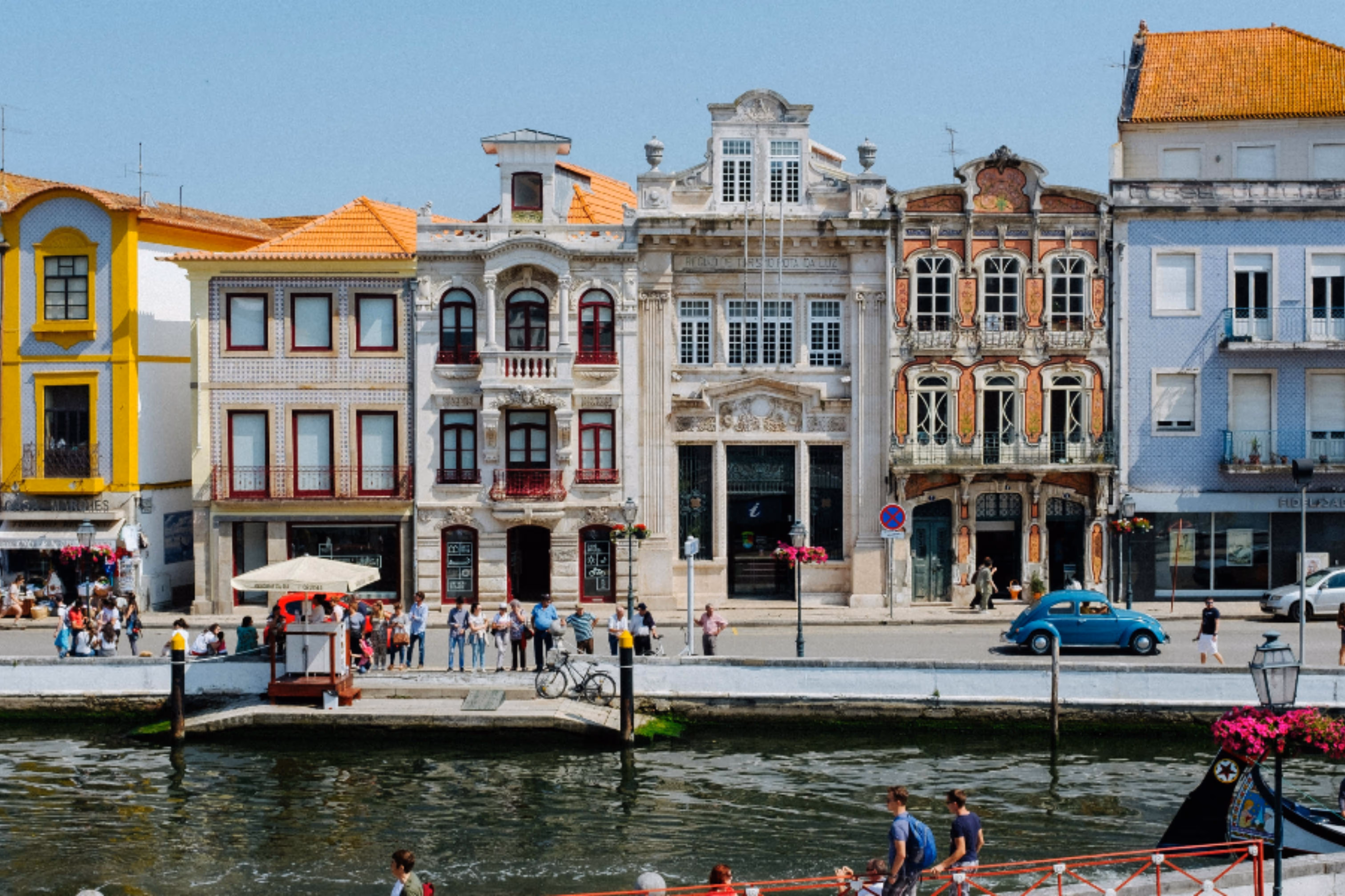 Colorful historic buildings with ornate facades along a waterfront promenade filled with people and a blue vintage car parked nearby.