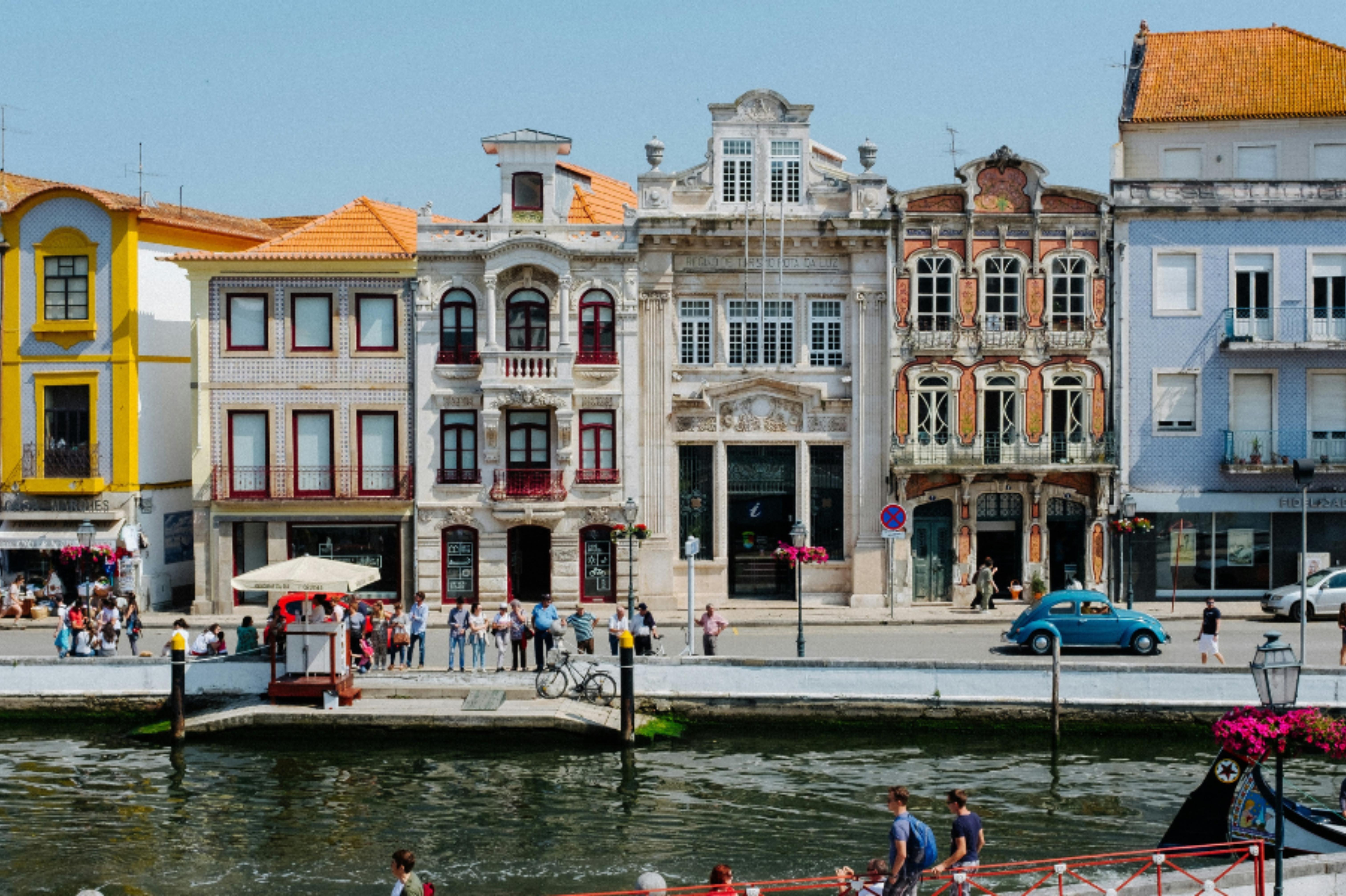 Colorful historic buildings with ornate facades along a waterfront promenade filled with people and a blue vintage car parked nearby.