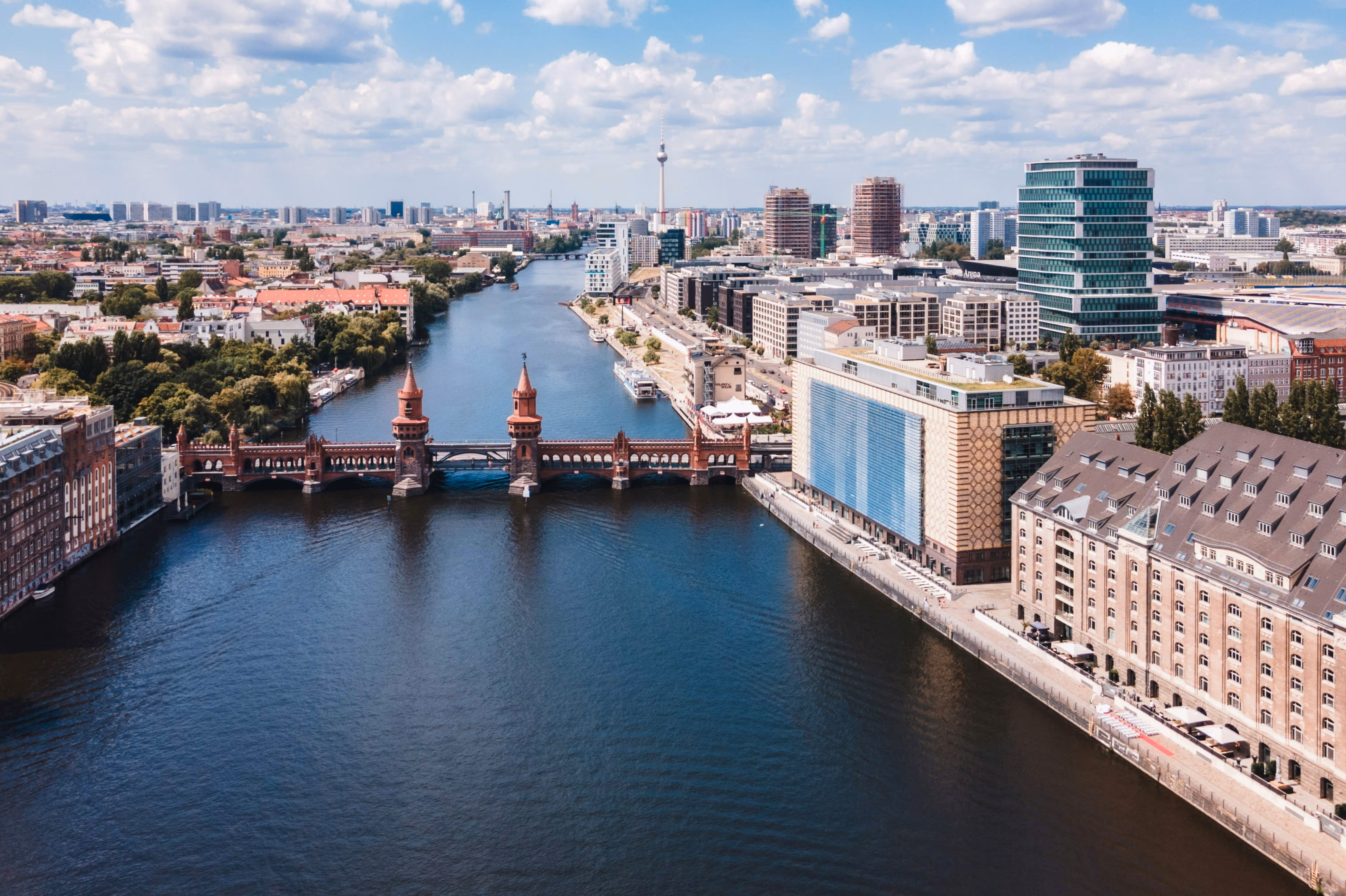 Aerial view of Berlin's Oberbaum Bridge over the Spree River with city buildings and TV tower under a partly cloudy sky.
