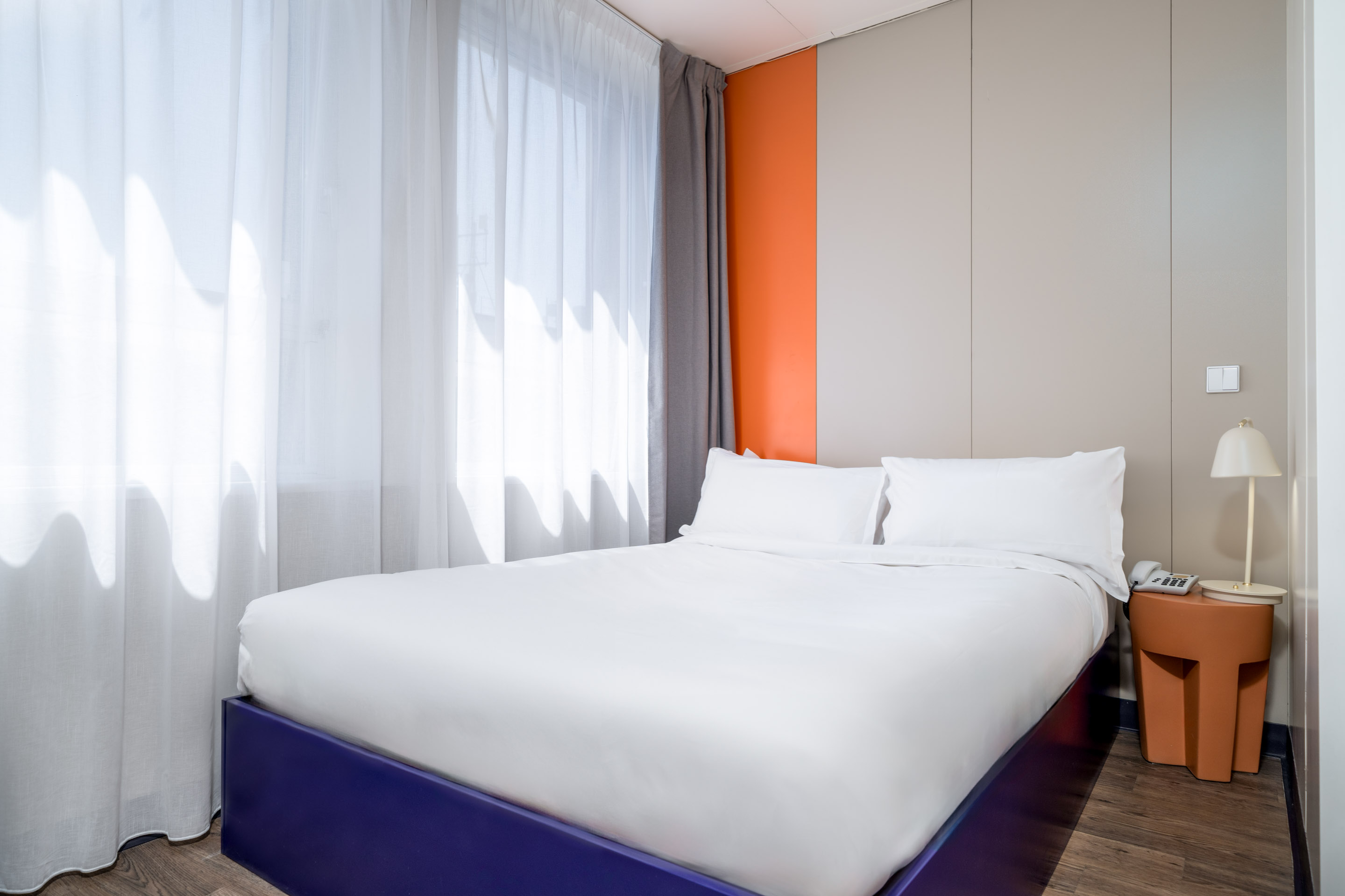 Modern hotel room with a neatly made bed featuring white linens, sheer white curtains, an orange accent wall, and a small bedside table with a lamp and telephone.