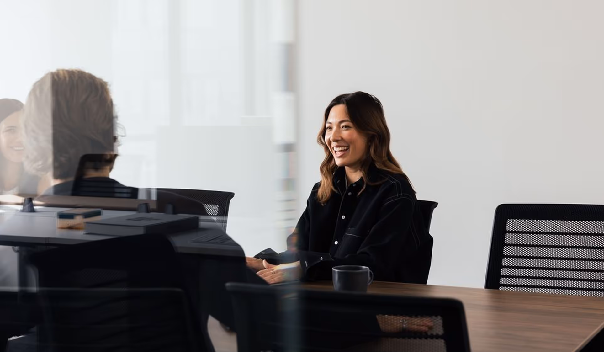 Two people seated at a conference table engaged in a friendly conversation, with one woman smiling brightly.