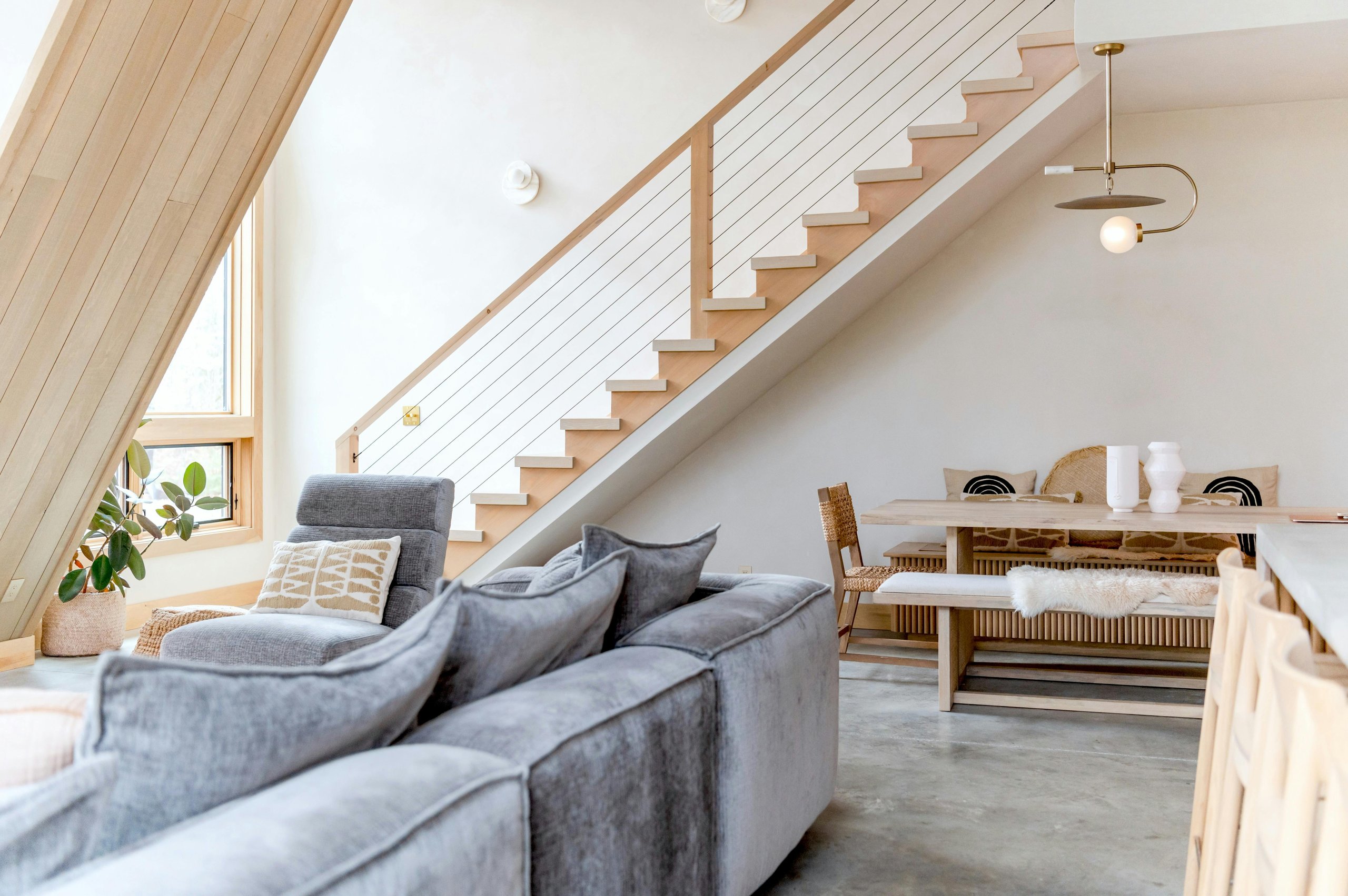 Modernes Wohnzimmer mit grauer Couch, einem Sessel mit beige-weißem Kissen, einer Pflanze neben einem großen Fenster und einer hölzernen Treppe mit Drahtgeländer.