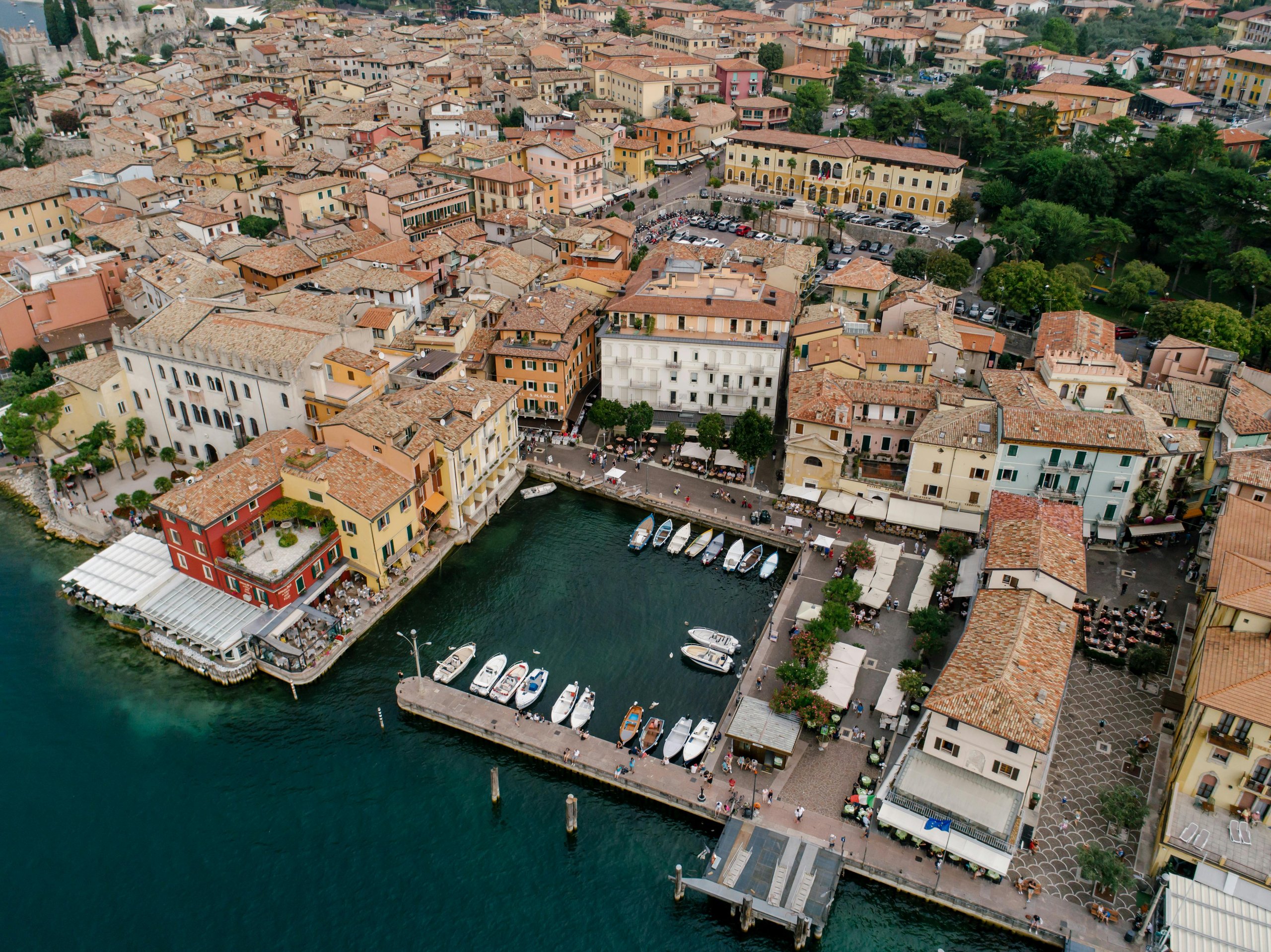 Vogelperspektive eines Hafens mit Anlegestellen und kleinen Booten vor einem pittoresken Küstendorf mit engen Gassen und terrakottafarbenen Dächern.