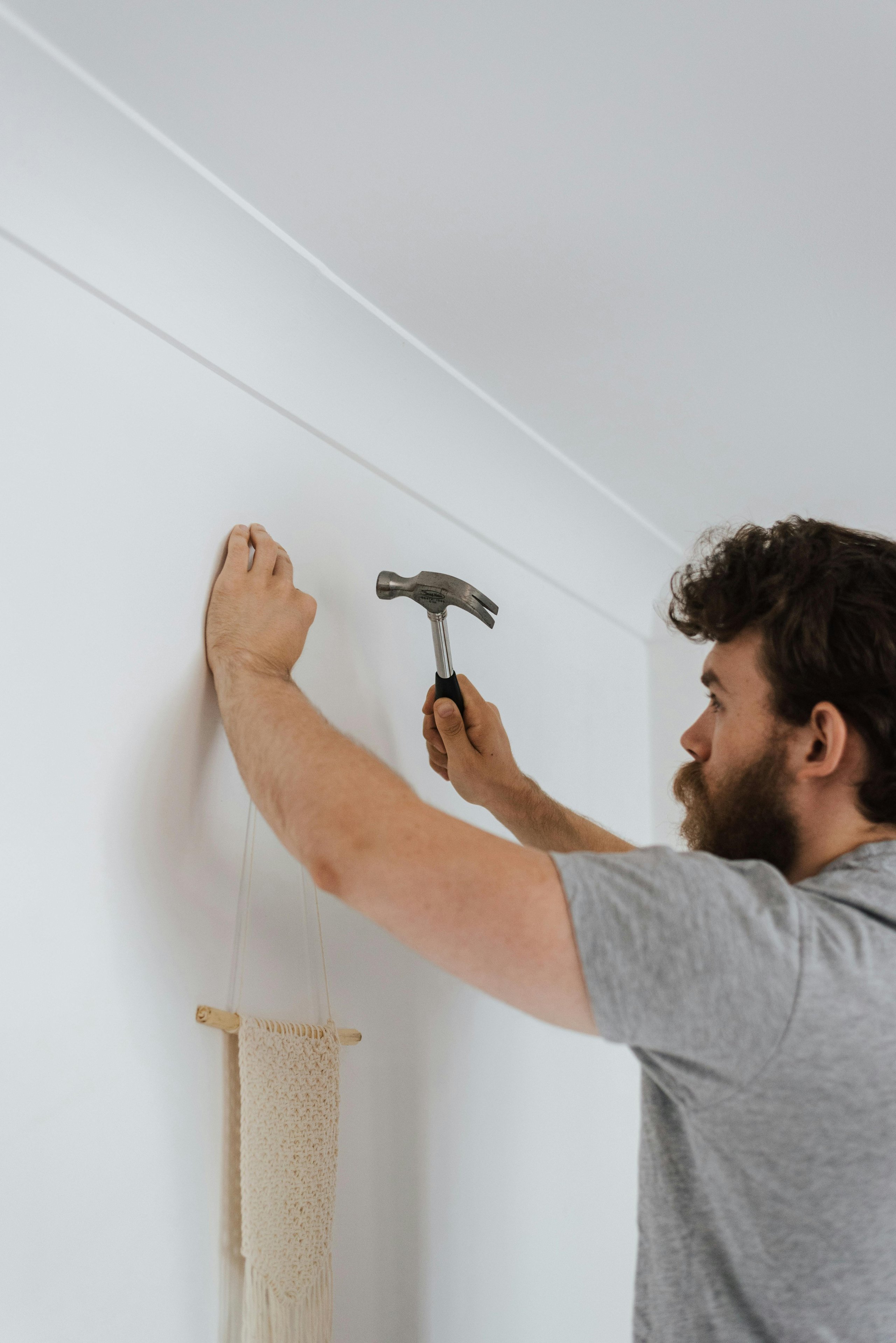 Ein Mann mit Bart schlägt mit einem Hammer einen Nagel in eine weiße Wand, um eine makramee Wanddekoration aufzuhängen.