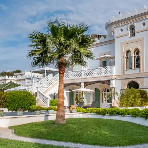 Palmier devant un bâtiment blanc de style méditerranéen avec des arcades, des balcons et une pelouse verte bien entretenue.