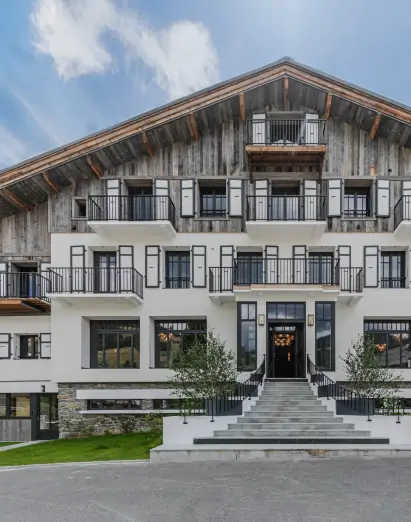 Vue de face de l'Écrin de Megève avec des murs supérieurs en bois, des balcons, de grandes fenêtres et un escalier central à l'entrée.