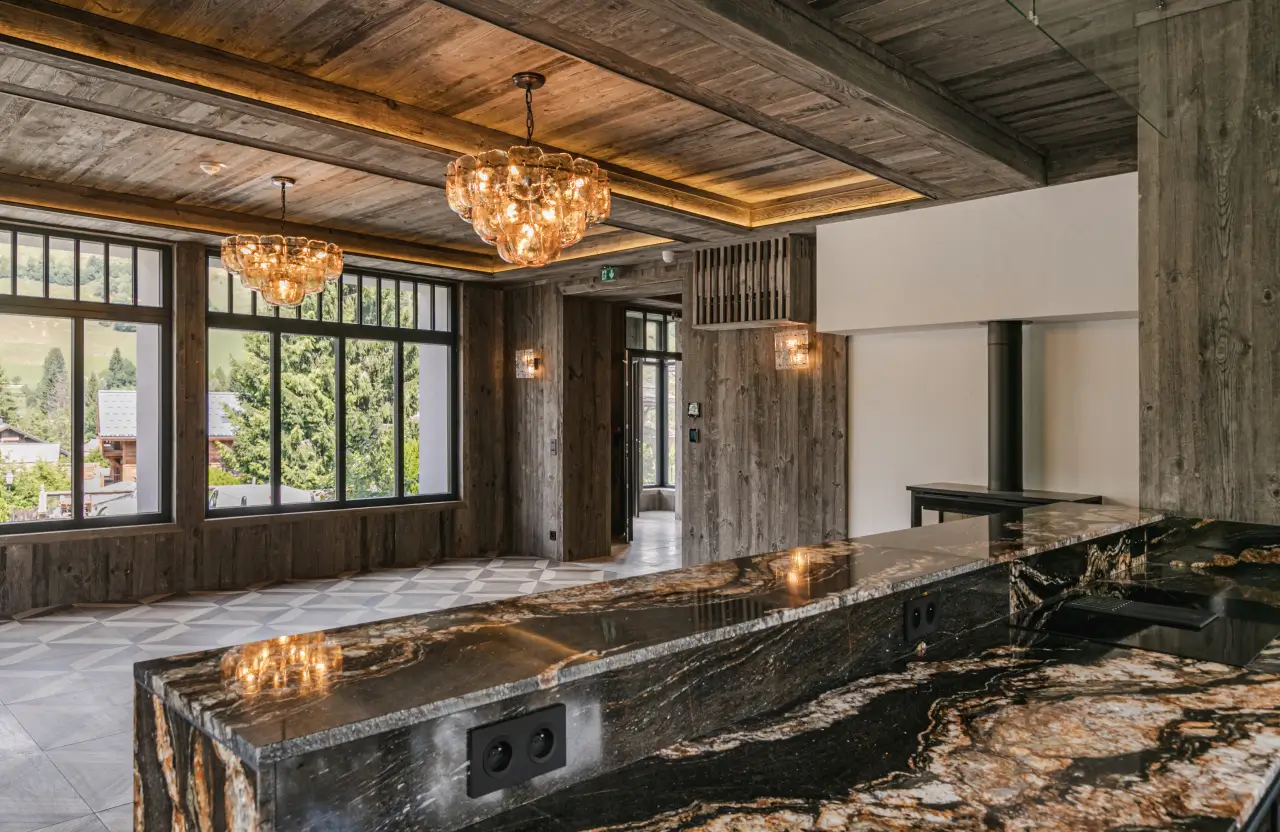 Cuisine moderne avec un grand plan de travail en marbre foncé, un plafond en bois, de grandes fenêtres et des suspensions lumineuses chaleureuses.