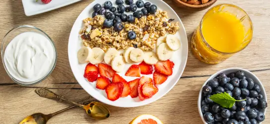 Bol de granola garni de myrtilles, de tranches de banane et de fraises sur une table en bois, avec à côté un verre de jus d'orange, un bol de myrtilles et un bol de yaourt.