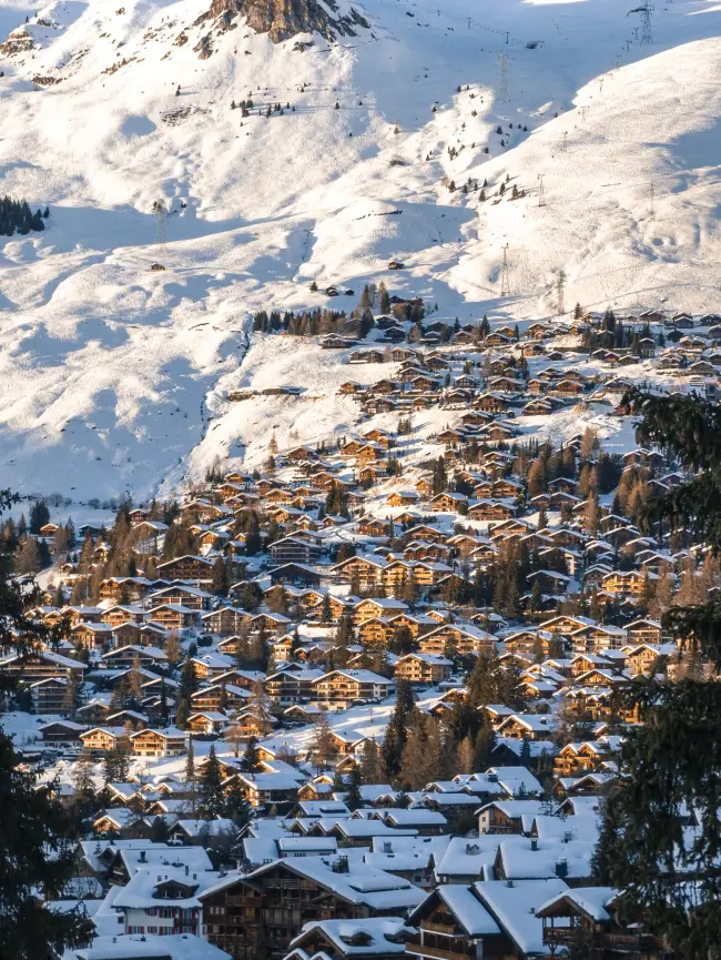 Village ensoleillé avec des maisons en bois recouvertes de neige à flanc de montagne, sous un sommet enneigé.