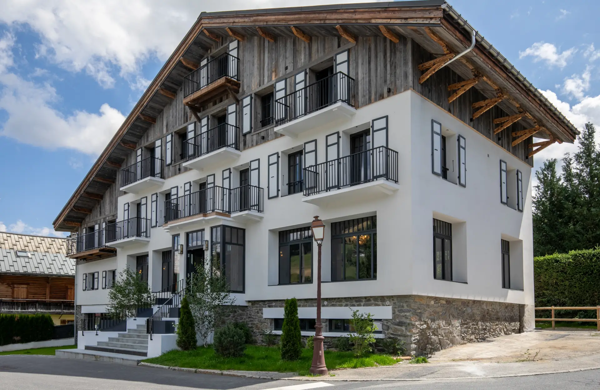 L'Écrin de Megève avec des murs blancs, des balustrades noires, une façade supérieure en bois et plusieurs balcons sous un ciel bleu parsemé de nuages.