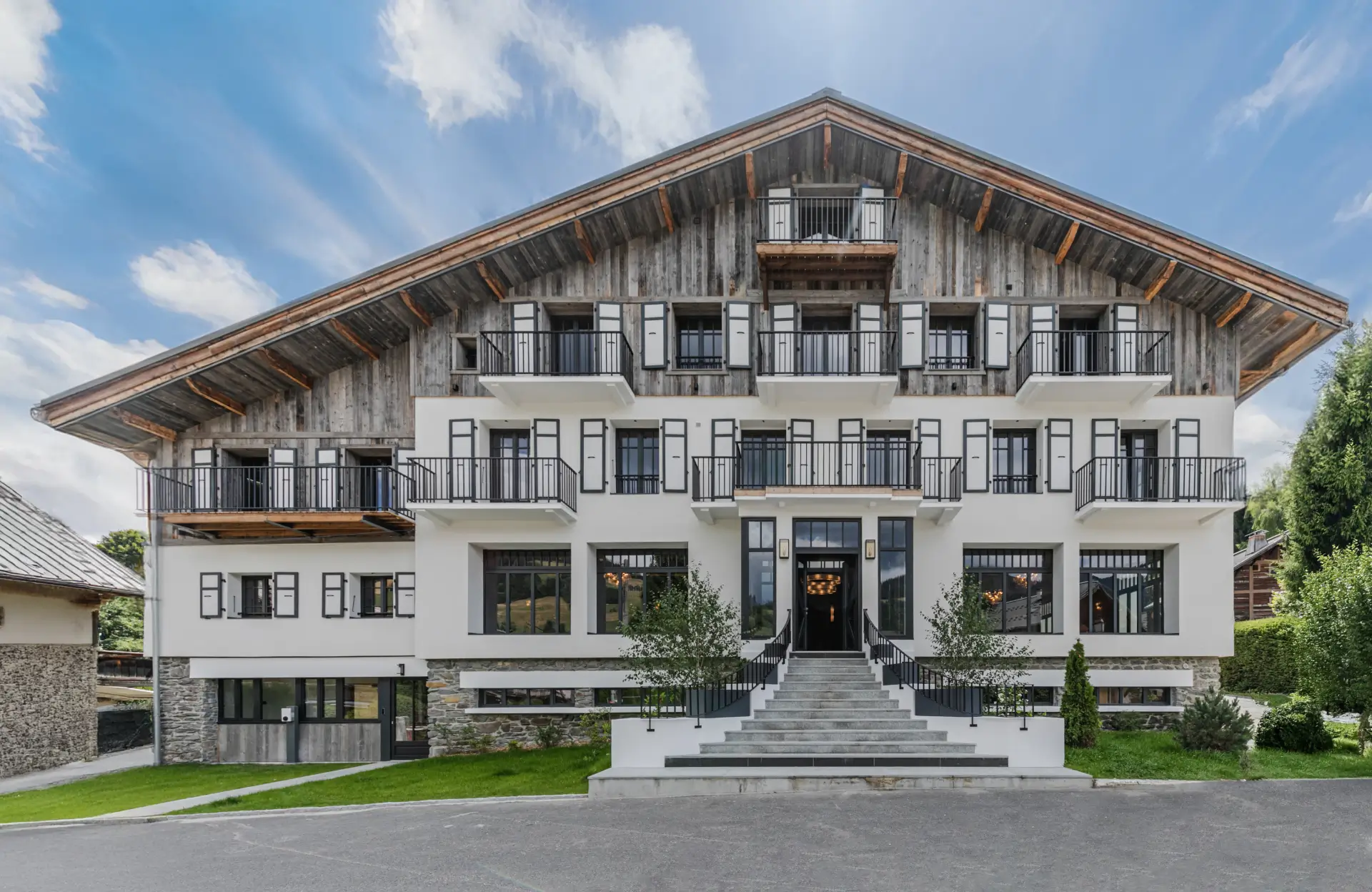 Vue de face de l'Écrin de Megève avec plusieurs balcons, une façade supérieure en bois et un large escalier d'entrée en pierre.