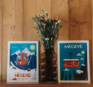 Deux affiches colorées de Megeve avec un téléphérique, placées sur une étagère en bois entourant un vase contenant des fleurs rouges et blanches.
