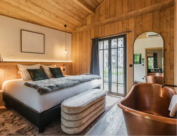Chambre moderne en bois avec un lit double, une baignoire cuivrée, un miroir arrondi et une porte vitrée donnant sur un balcon.