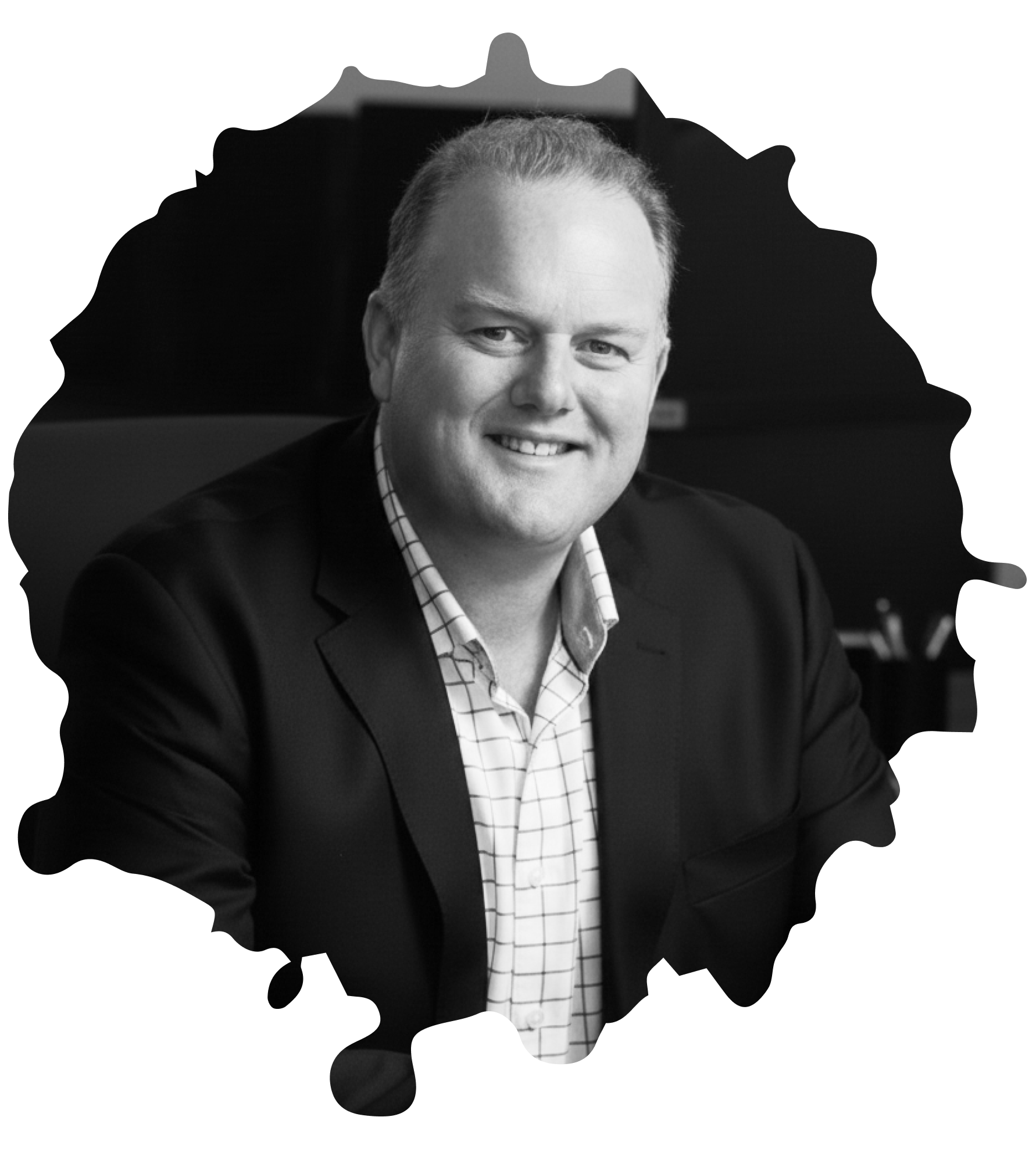 Smiling man in a checked shirt and dark blazer sitting indoors.