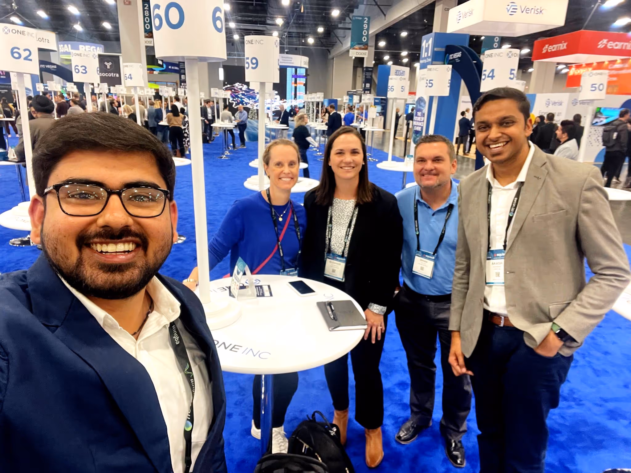 Five professionals smiling and posing for a selfie at a conference with numbered signs and booths in the background.