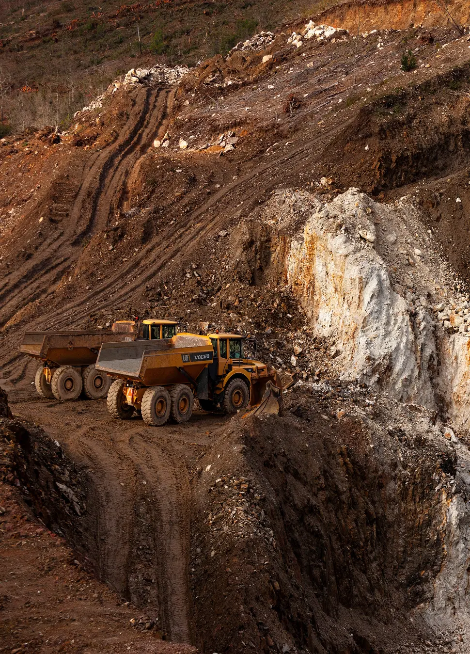 Two yellow Volvo dump trucks parked on a dirt mining site with rocky terrain.