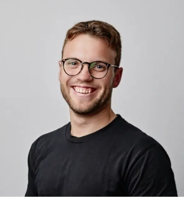 Smiling young man with glasses and short brown hair wearing a black shirt against a gray background.