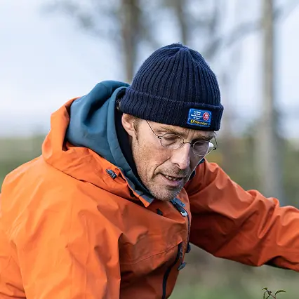 Man wearing glasses, a navy knit beanie, and an orange jacket looking downward outdoors.