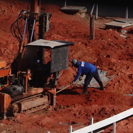 Worker wearing a blue shirt and helmet shoveling red dirt near construction machinery on a site.