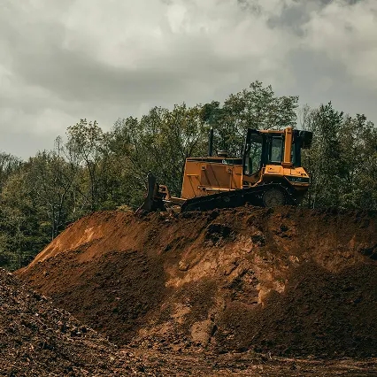 Yellow bulldozer parked on top of a large mound of dirt with trees and cloudy sky in the background.