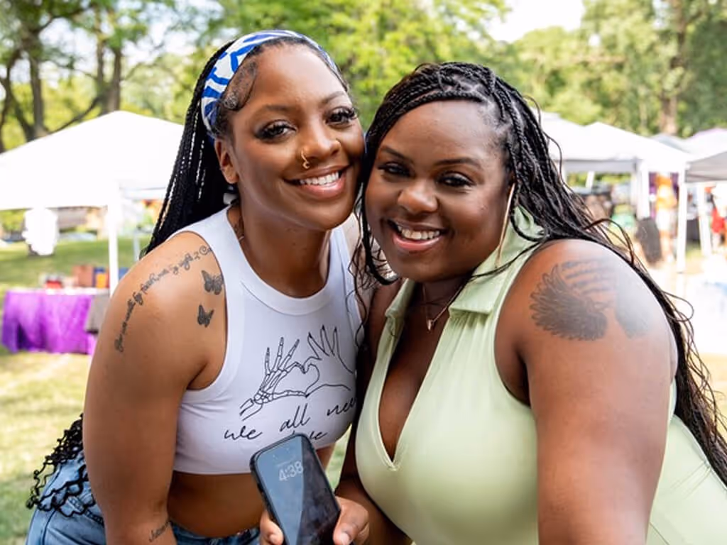Two smiling women with braided hair and tattoos posing closely together outdoors at a sunny event.
