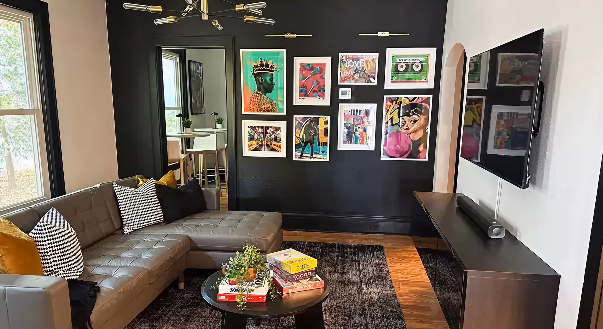 Modern living room with gray sectional sofa, black and white and mustard pillows, round black coffee table with board games and plants, black accent wall with colorful framed artwork, and a wall-mounted TV.