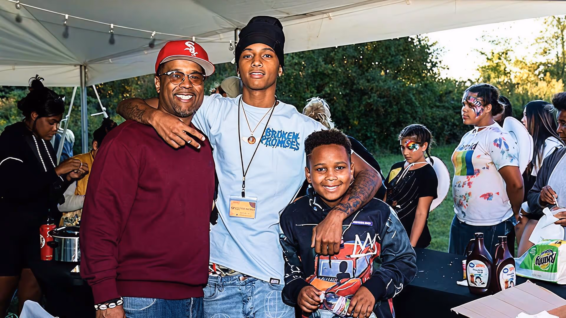 Three males smiling under a tent at an outdoor event, with people in face paint and wings in the background.