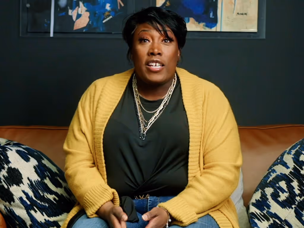 Person wearing a yellow cardigan and layered necklaces sitting on a couch with patterned pillows.
