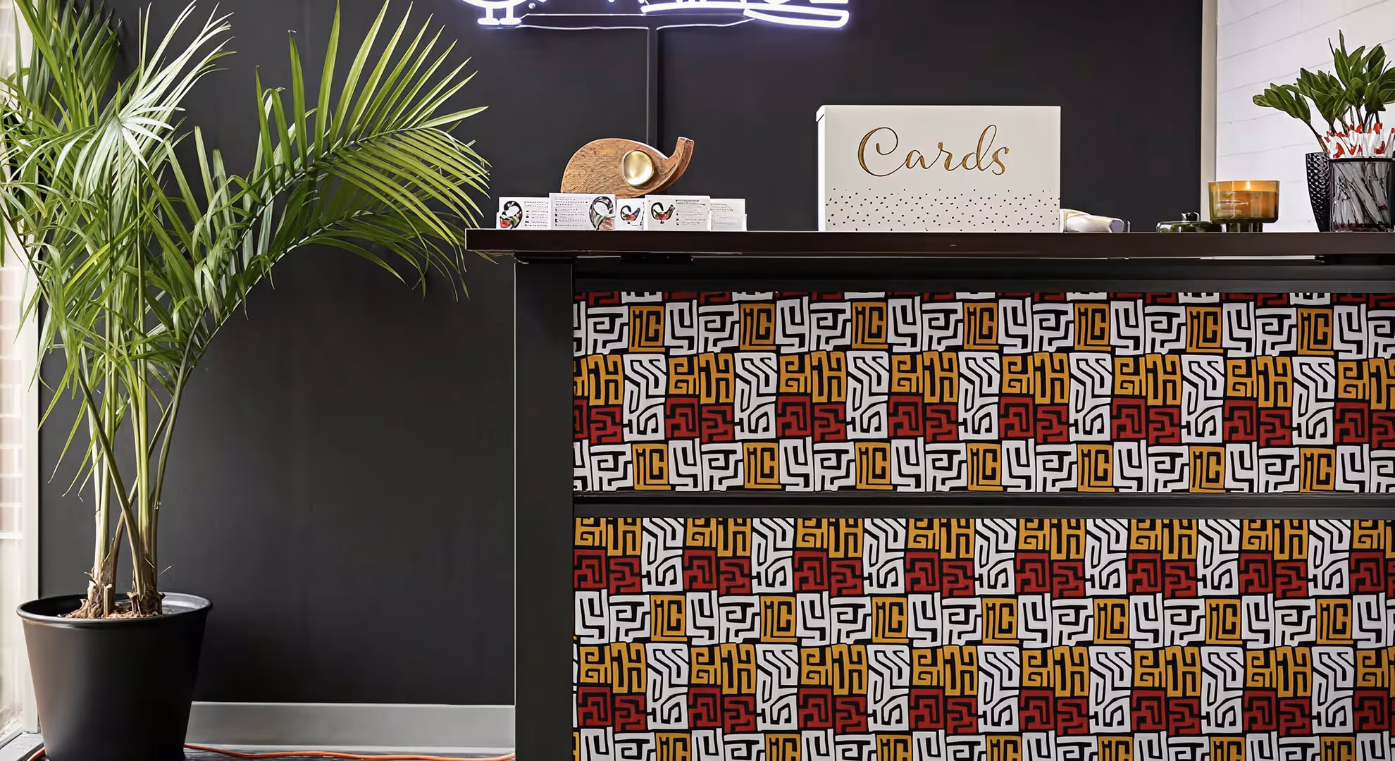 Reception desk with a multicolored geometric patterned front, a large potted palm plant on the left, and a card box on the black tabletop.