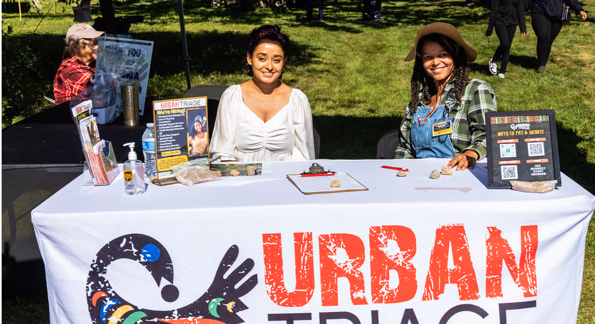 Two women smiling while sitting behind an Urban Triage information and hiring table outdoors on a sunny day.
