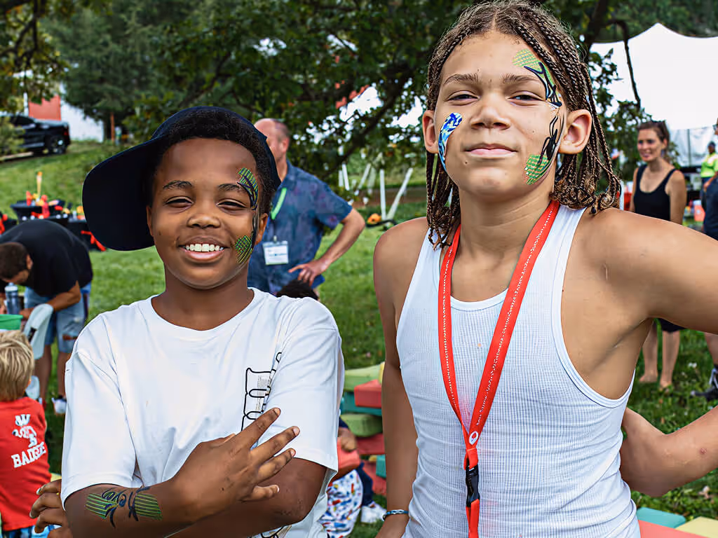 Two boys with face paint posing and smiling outdoors at a festival with people and trees in the background.