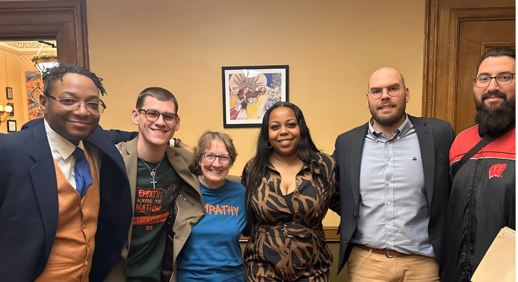 Six diverse adults smiling together in an indoor room with beige walls and framed artwork.
