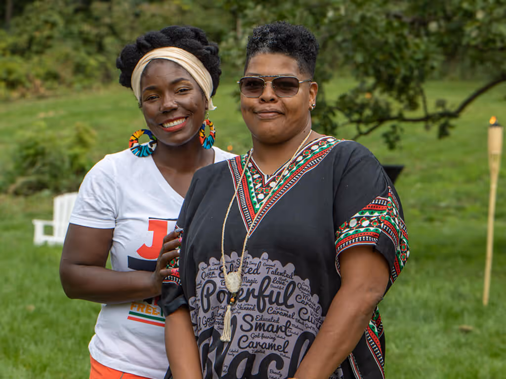 Two smiling Black women standing outdoors on green grass with trees in the background.