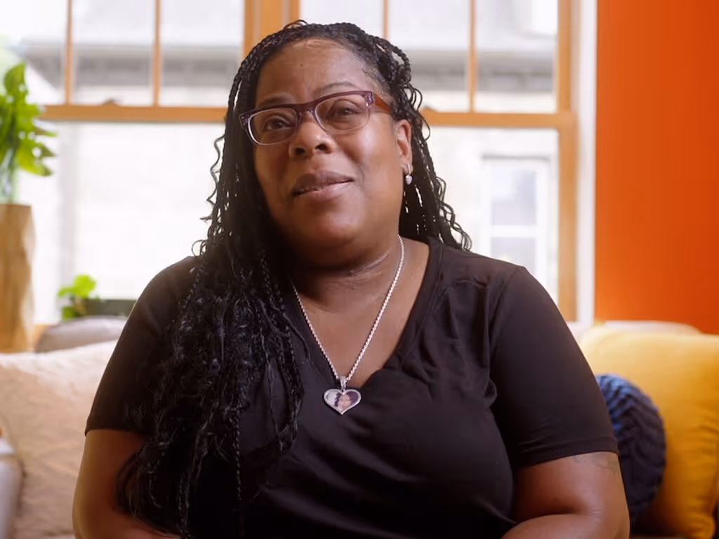 Smiling woman with glasses and braided hair wearing a black top and heart-shaped necklace sitting on a couch in a bright room.