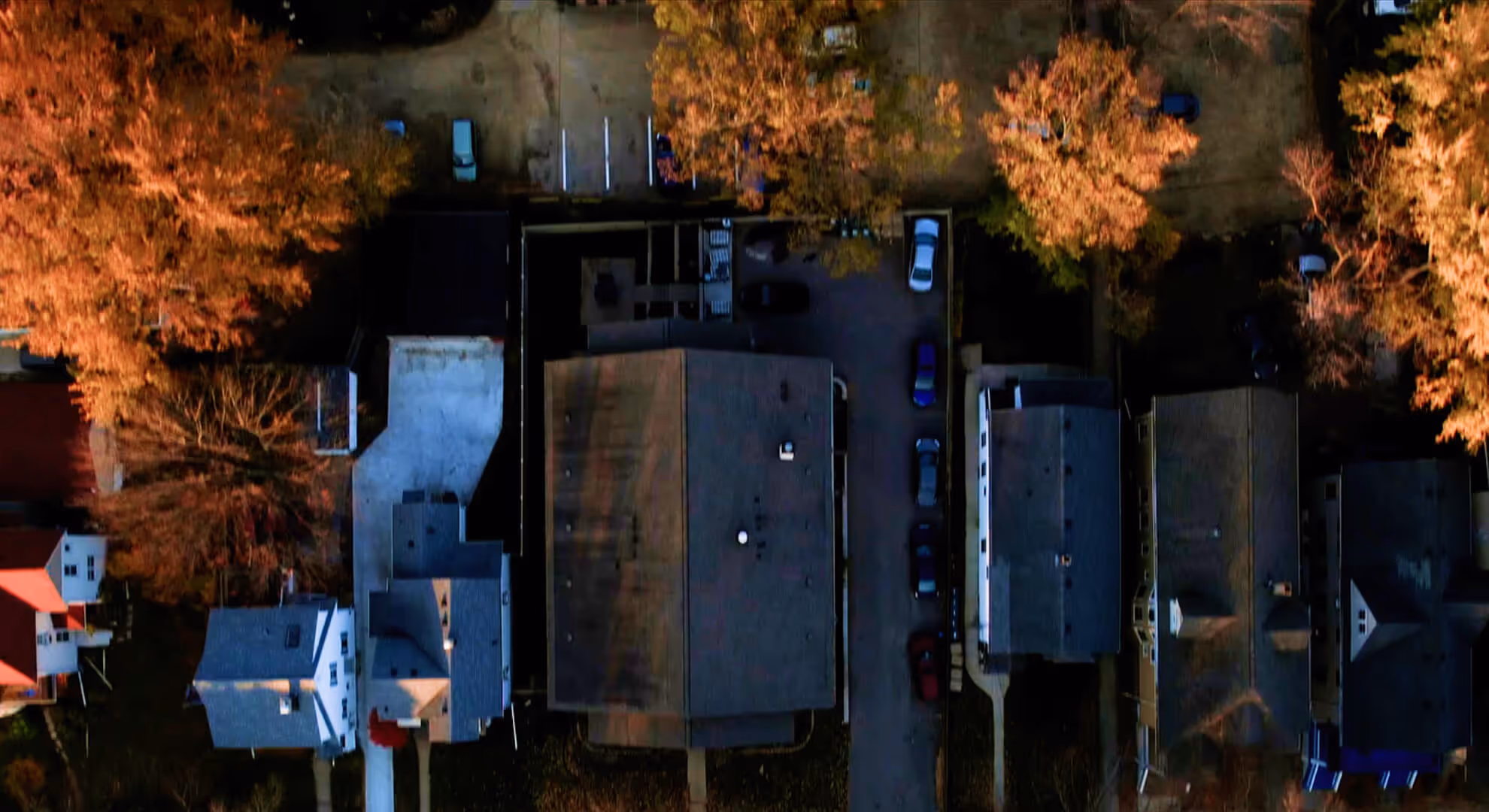 Aerial view of residential houses surrounded by autumn-colored trees and parked cars along the street.