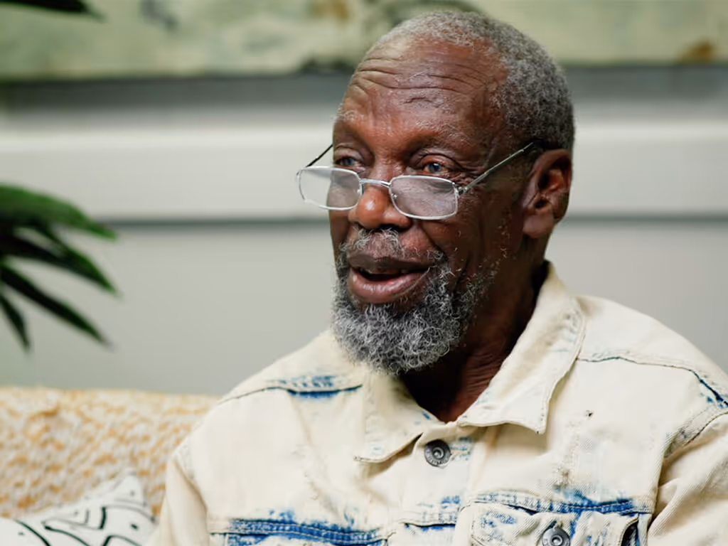 Smiling elderly man with gray beard wearing glasses and a light denim jacket indoors.