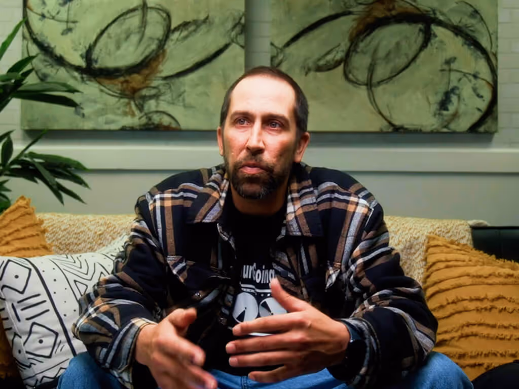 A man with a beard wearing a plaid shirt sits on a couch with decorative pillows, gesturing with his hands.