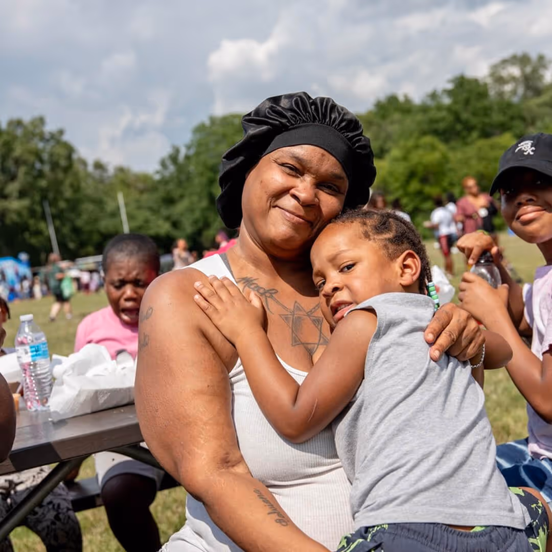 A smiling woman wearing a black bonnet hugs a young child with braided hair outdoors at a park gathering.