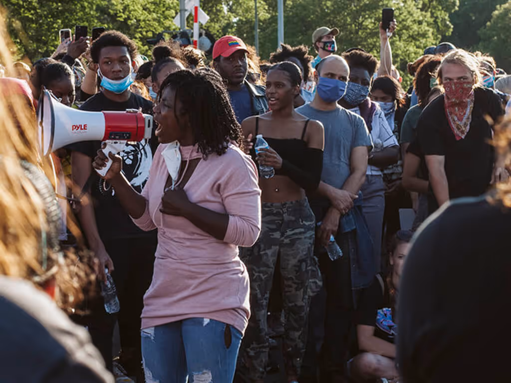Woman speaking through a megaphone in front of a diverse crowd, some wearing masks, outdoors with trees in the background.