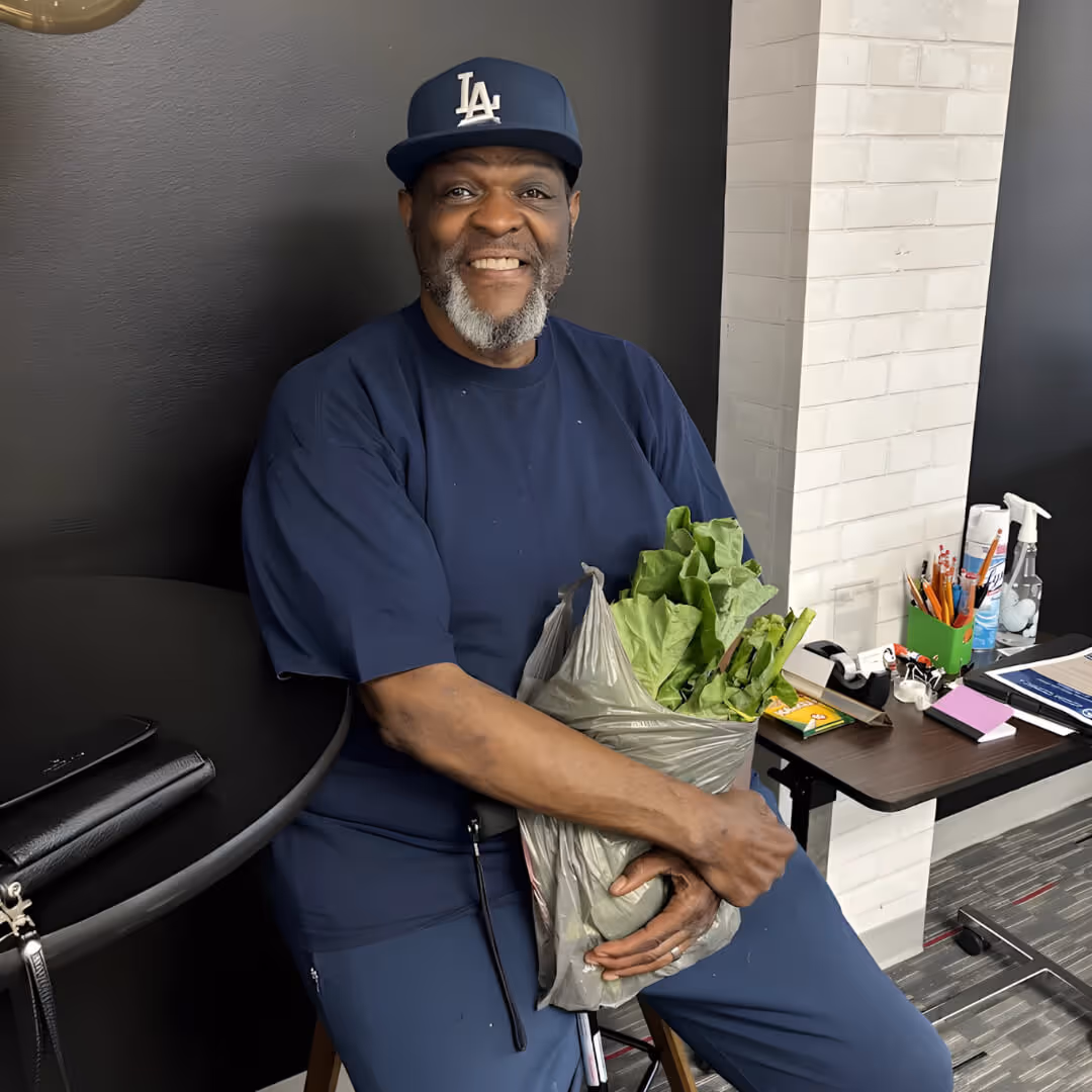 Smiling man in navy blue attire and LA cap holding a plastic bag filled with leafy green vegetables while seated indoors.