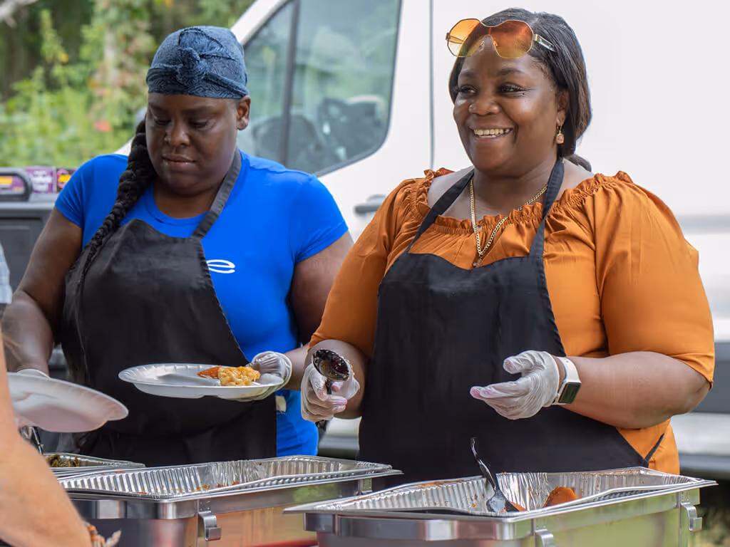 Two women in aprons serving food at an outdoor buffet, one smiling while holding a bottle and the other focused on a plate.