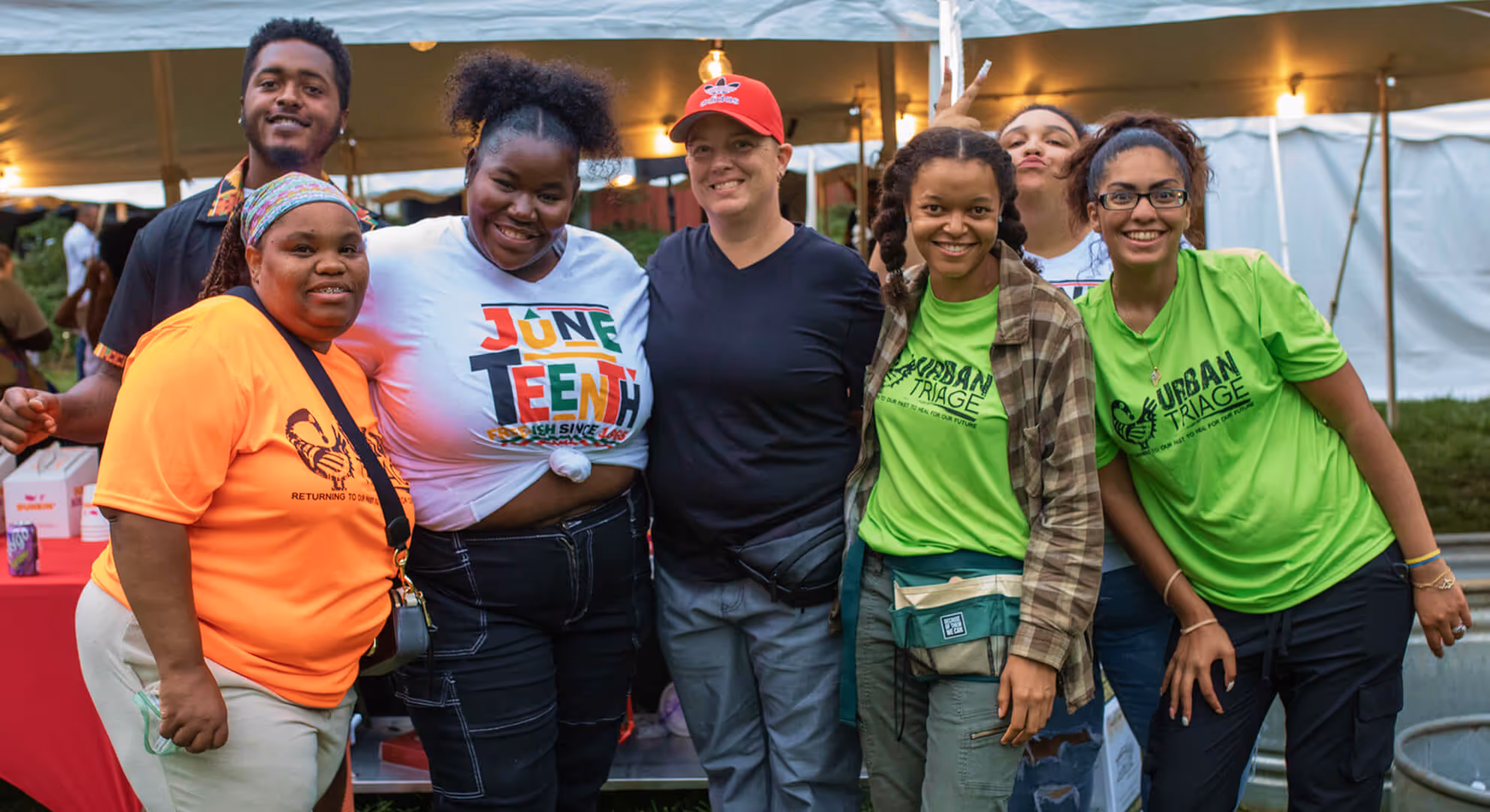 Group of seven diverse people smiling and posing together under a tent at an outdoor event.