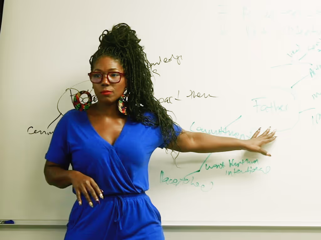 Woman in blue dress with glasses and large earrings pointing at whiteboard with handwritten notes.