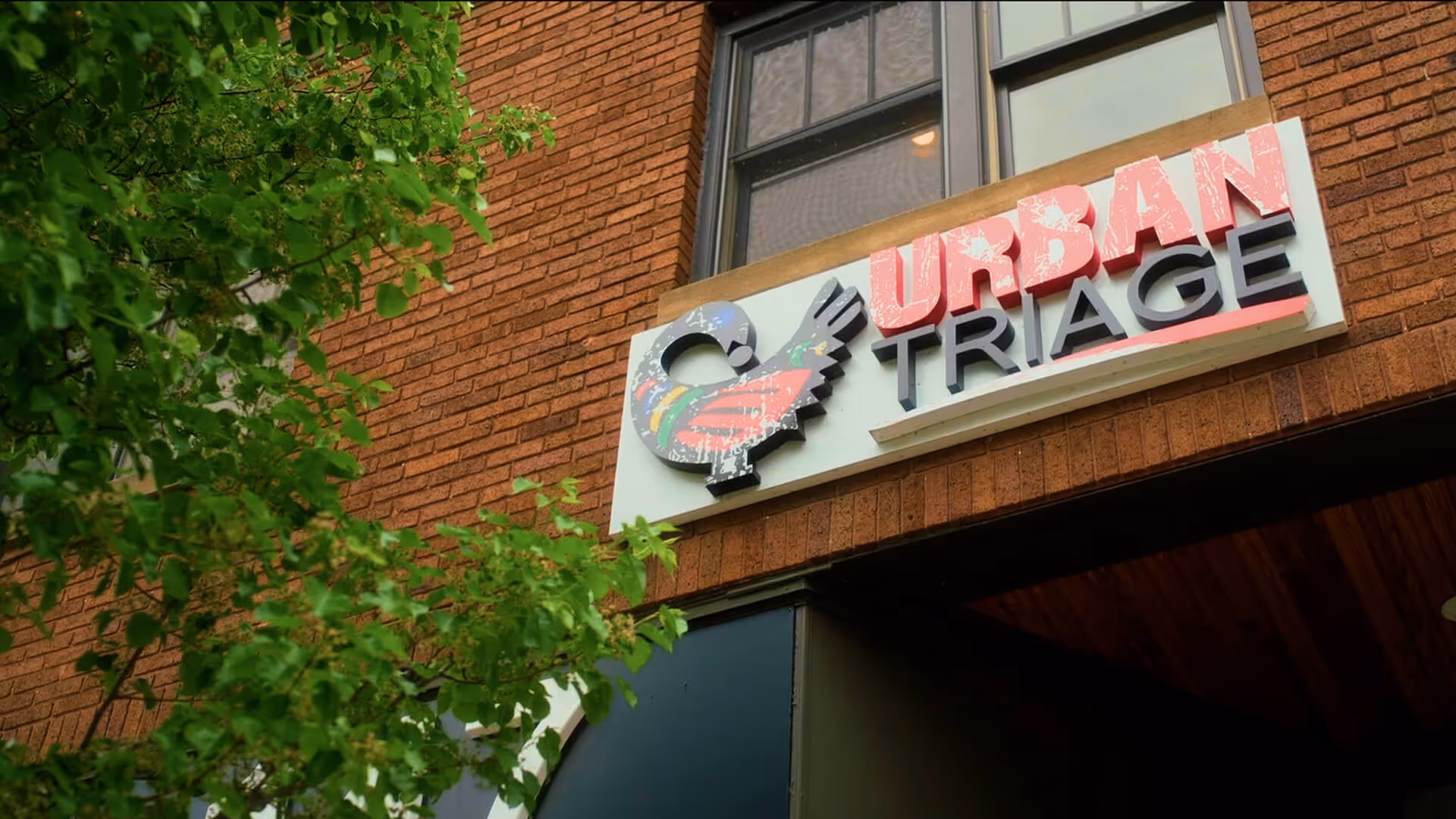 Sign on a brick building reading 'Urban Triage' with a colorful bird logo, partially obscured by green leaves.