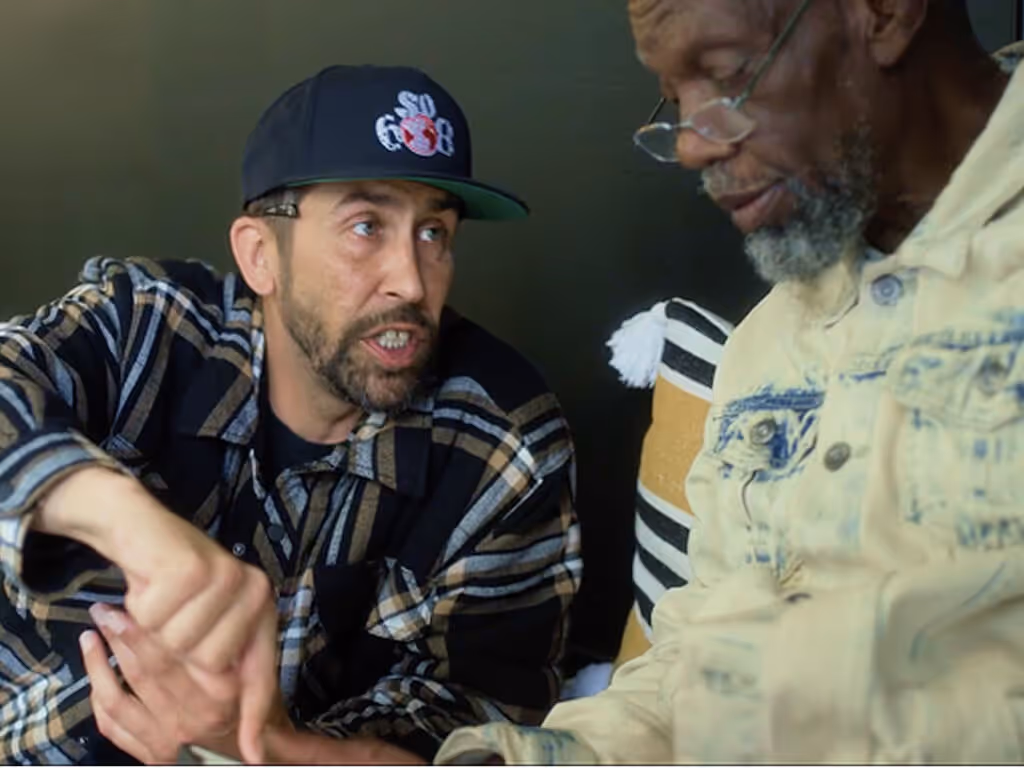 Two men sitting closely and engaged in a serious conversation, one wearing a black and beige plaid jacket and a cap, the other in a light denim jacket with glasses.