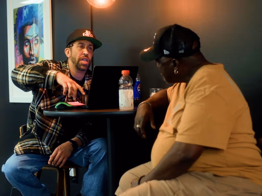 Two men sitting at a small round table engaged in conversation, one using a laptop and the other listening attentively.