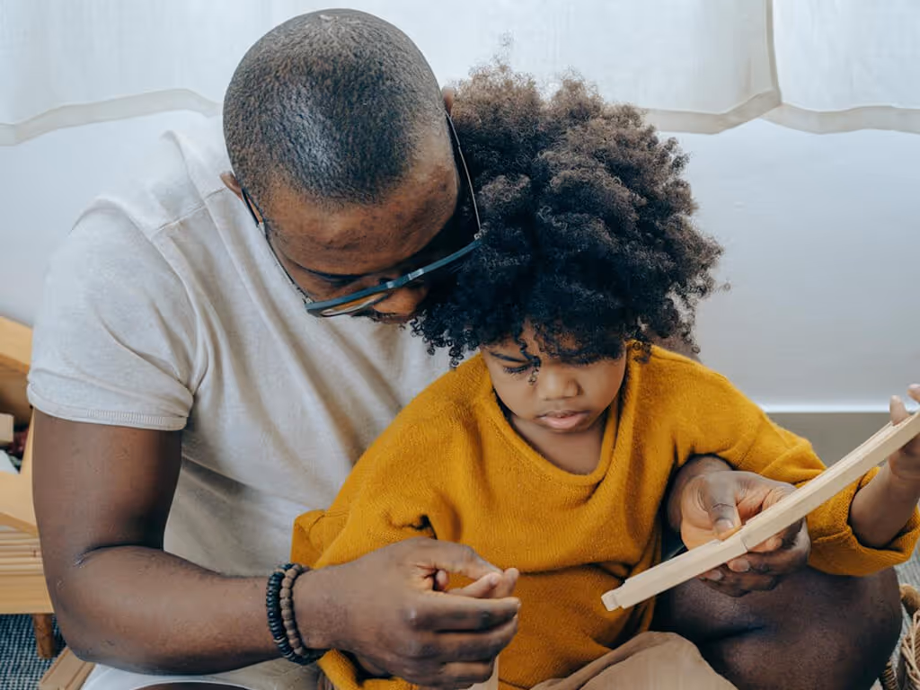 Father and young child closely reading a book together near a window with curtains.