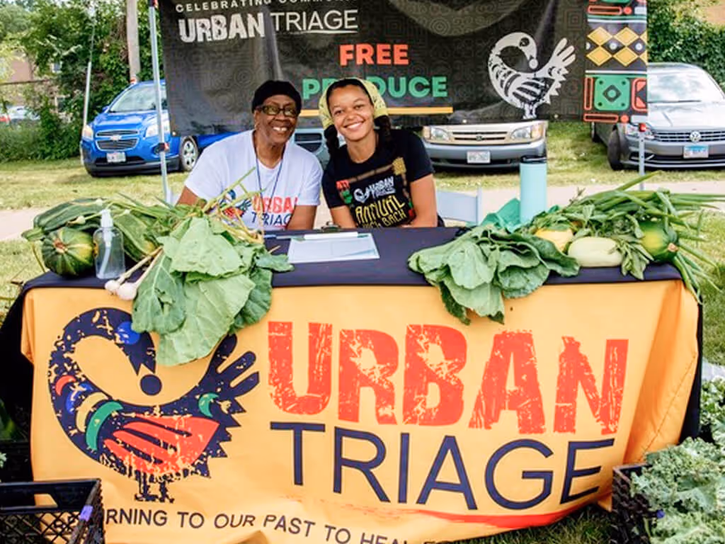 Two smiling women sitting behind a table with fresh produce and an Urban Triage banner offering free produce.