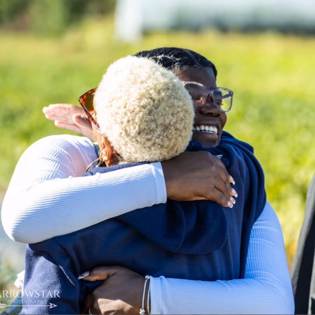 Two people warmly hugging outdoors, one smiling with glasses and the other with curly blonde hair and sunglasses.
