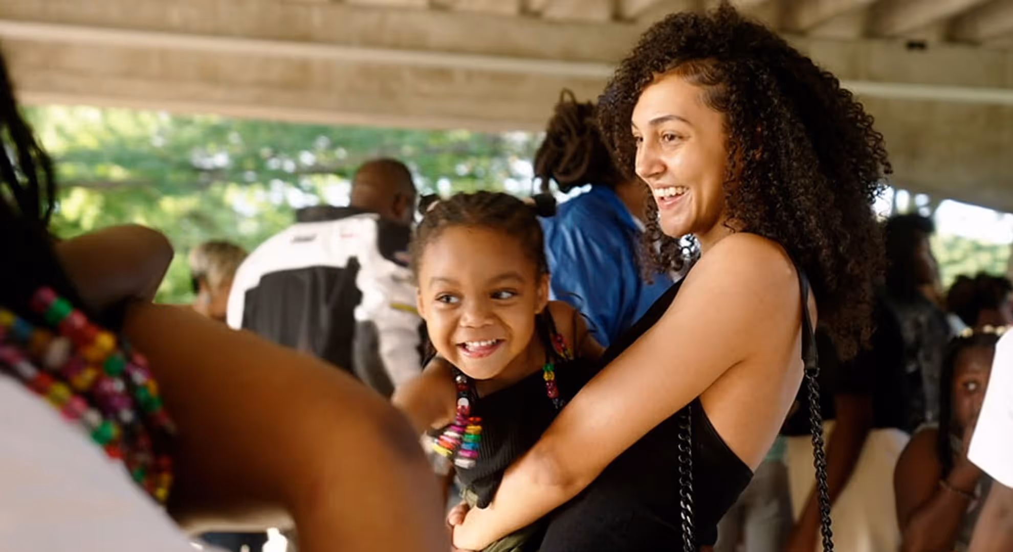 Smiling woman with curly hair holding a happy young girl with beaded braids at an outdoor gathering.
